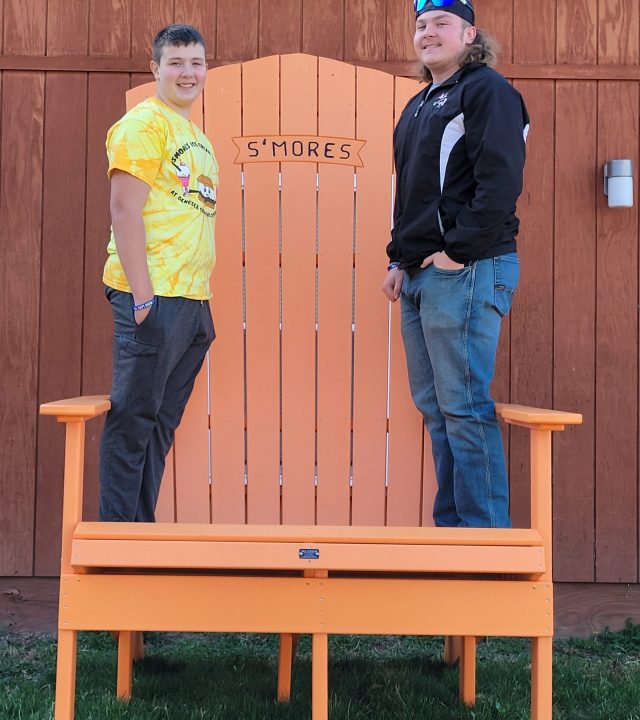 Giant S’Mores Chair photo at the campground – fun photo spot for guests Two guests standing on an oversized orange Adirondack chair labeled “S’Mores” at a campground